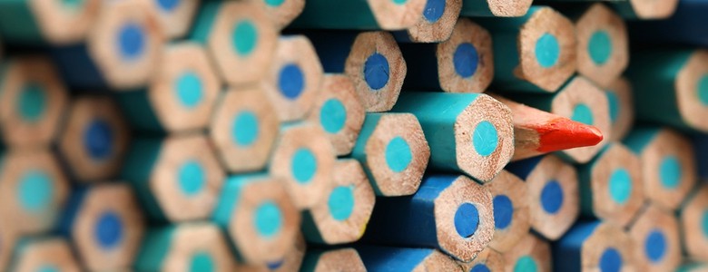 A single orange pencil facing outwards among a wall of blue pencils facing the other way